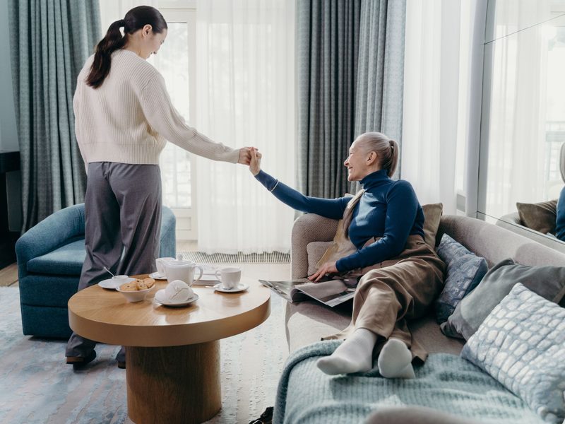 A caregiver helping an older woman up from a sofa in a bright living room